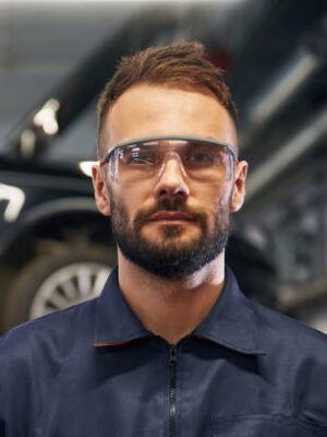 Looking at the camera. car repairman is in the garage with automobile.