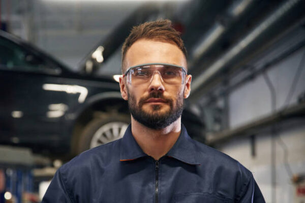 Looking at the camera. car repairman is in the garage with automobile.